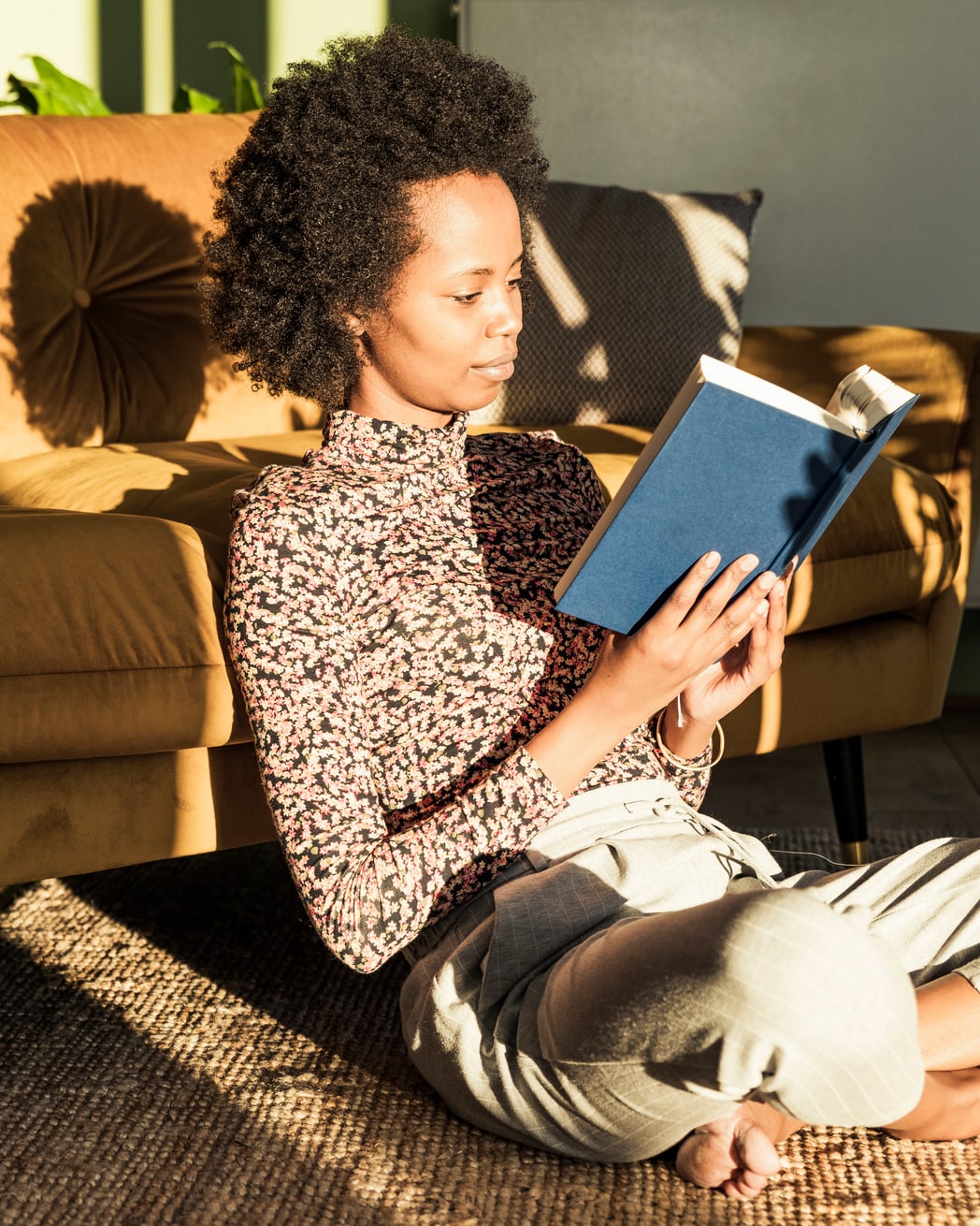 Une femme est assise par terre, appuyée contre un canapé en cuir. Elle lit un livre dans une pièce ensoleillée.
