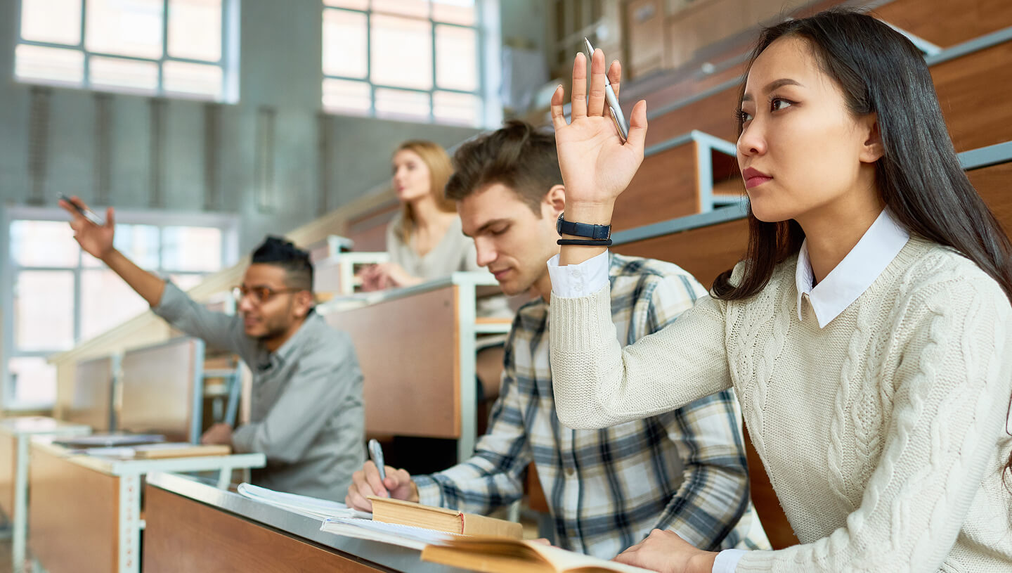 Une salle de classe universitaire avec des étudiants assis à des bureaux, tandis qu’une étudiante au premier plan lève la main.